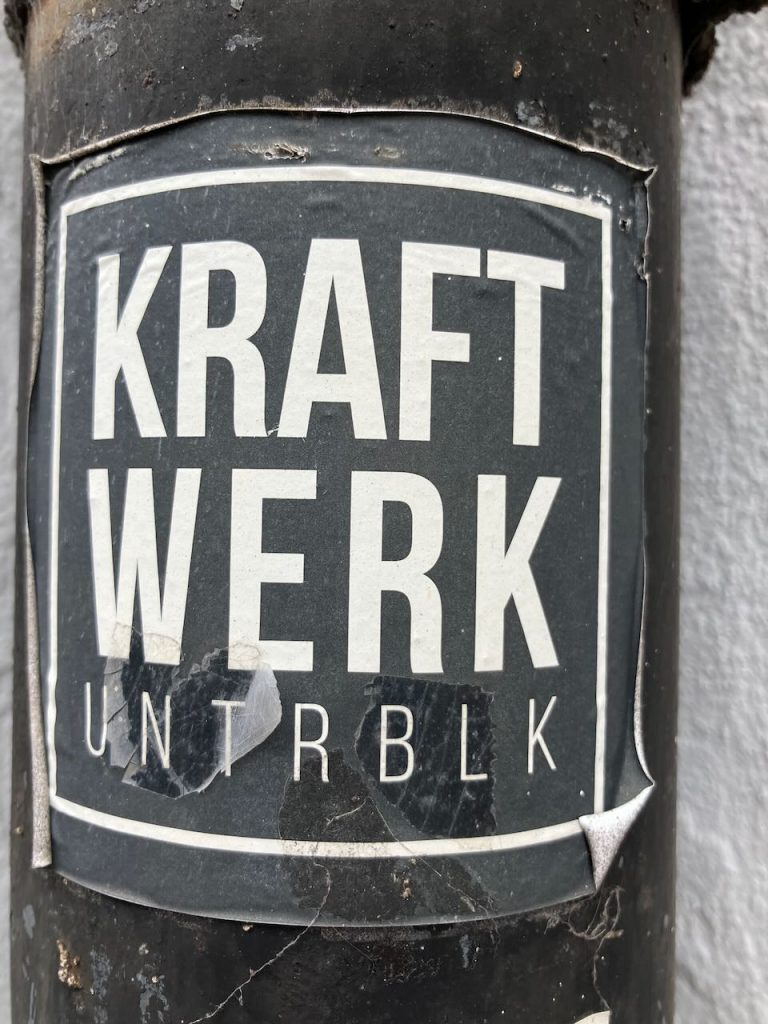 Bildbeschreibung: Ein rechteckiger, schwarz-weißer Sticker mit der Aufschrift „KRAFT WERK UNTRBLK“ in großen, klaren Buchstaben. Der Aufkleber ist leicht abgenutzt und auf einer dunklen Oberfläche angebracht.