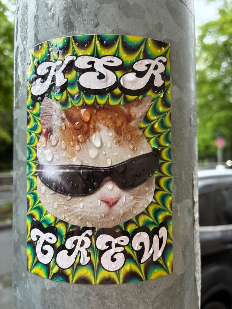 Sticker mit psychedelischem, gelb-grünem Muster. In der Mitte ist ein Katzenkopf mit Sonnenbrille zu sehen. Oben steht „KSR“, unten „CREW“ in weißer, verschnörkelter Schrift.