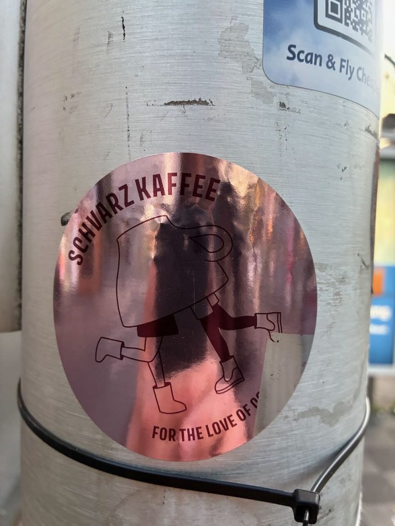 Runder, glänzender Sticker in Roségold. Darauf die Aufschrift „SCHWARZ KAFFEE“ und „FOR THE LOVE OF COFFEE“. In der Mitte eine Zeichnung einer laufenden Kaffeetasse mit Beinen und Stiefeln.