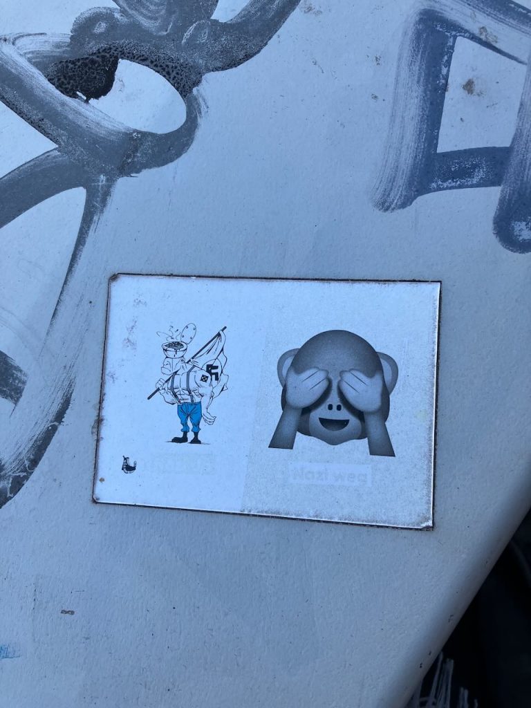 Links eine gezeichnete Figur in weißer Robe mit Kapuze, die ein Hakenkreuz trägt. Rechts daneben ein Emoji-Affe, der sich die Augen zuhält. Unten steht blass: „Nazi weg“.