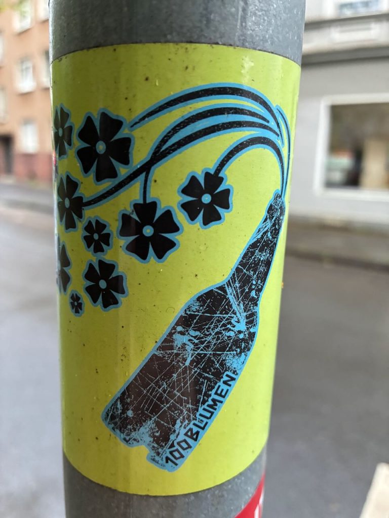 Ein gelber Sticker zeigt eine schwarze, zerbrochene Flasche, aus der stilisierte blaue Blumen wachsen. Auf der Flasche steht „100 BLUMEN“.
