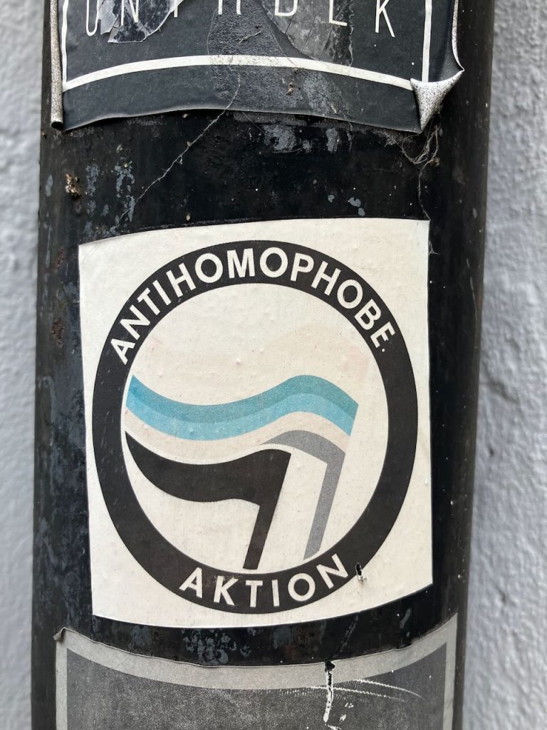 Bildbeschreibung: Ein runder Sticker mit schwarzem Rand und der Aufschrift „ANTIHOMOPHOBE AKTION“. In der Mitte sind zwei stilisierte Fahnen in Schwarz und Hellblau zu sehen. Das Motiv steht für Toleranz und Vielfalt.
