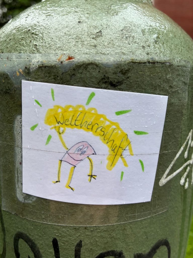 Handgezeichneter Sticker mit einem kleinen, rosa-gelben Männchen, das einen großen, gelben Schriftzug „Weltherrschaft“ über dem Kopf hält. Der Hintergrund ist weiß.