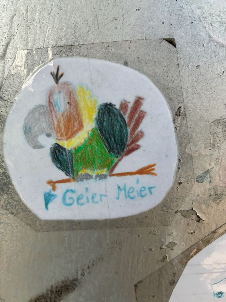 Handgezeichneter, bunter Geier auf einem Ast. Darunter steht „Geier Meier“.
