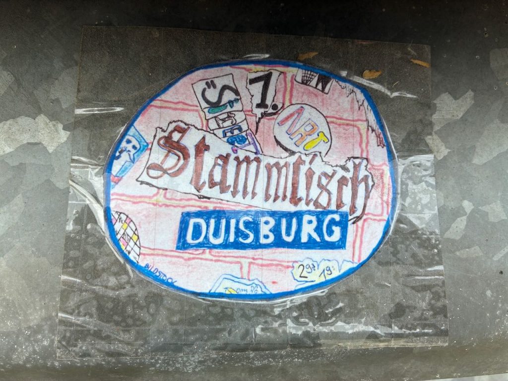 Ein runder Sticker hinter einer durchsichtigen Plastikfolie. Mit Buntstiften steht darauf geschrieben: 1. Streetart-Art-Stammtisch. Die einzelnen Wörter sind als farbige Sticker auf einer Ziegelmauer gezeichnet.