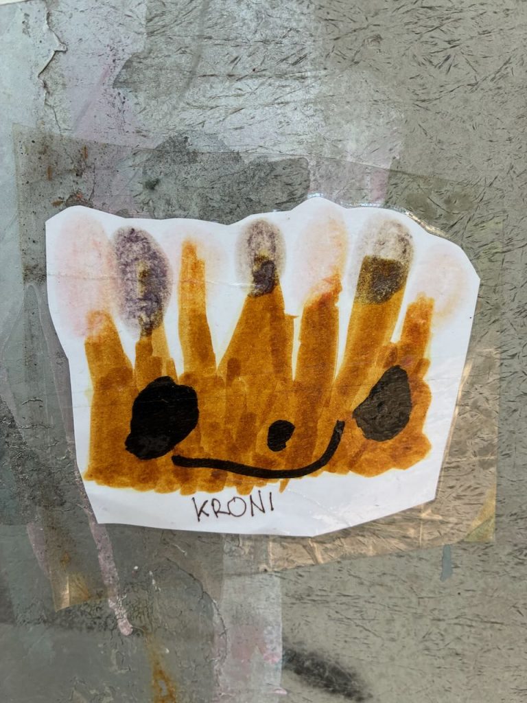 Ein stilisierter, brauner Abdruck in Form einer Krone mit schwarzen Punkten und einem schwarzen Mund, darunter steht „KRONI“.