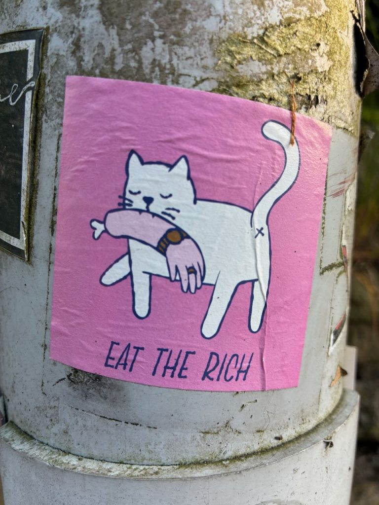 Eine weiße Katze auf pinkem Hintergrund hält einen menschlichen Arm im Maul. Darunter steht „EAT THE RICH“.