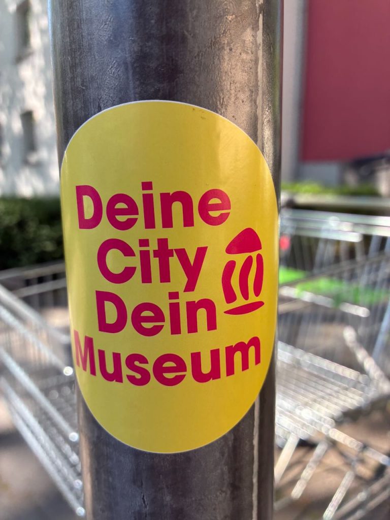 Ein gelber Sticker mit der Aufschrift „Deine City Dein Museum“ in Pink und einem kleinen Symbol, auf einer Metallstange mit Einkaufswagen im Hintergrund.