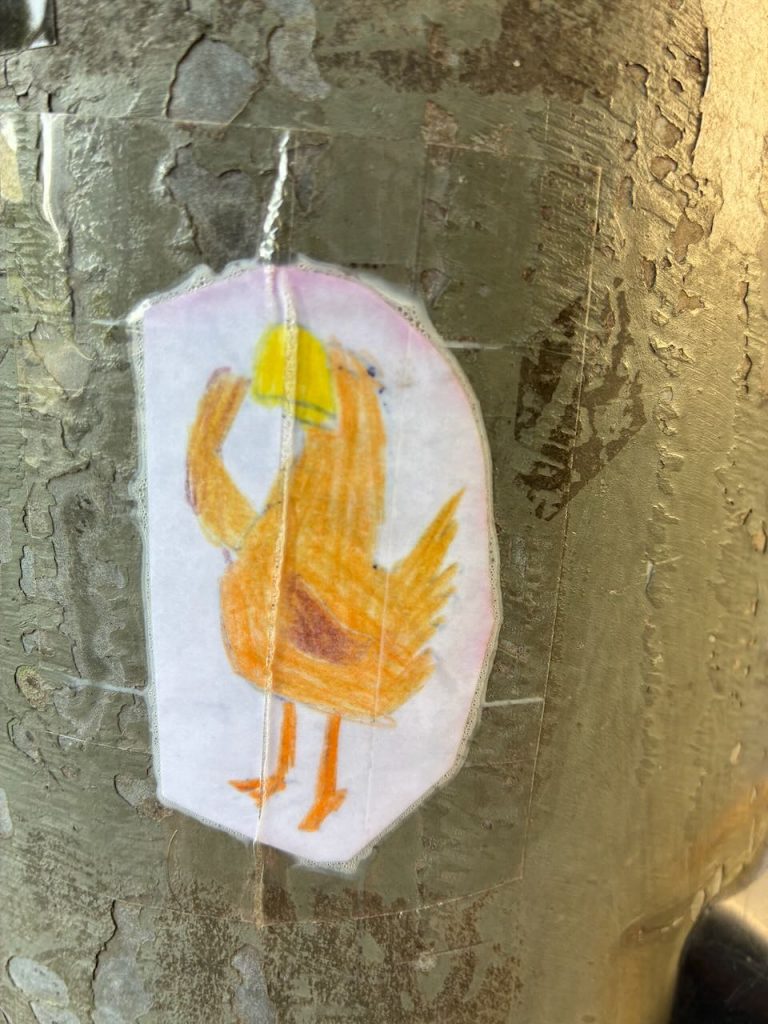 Auf einem silbernen Metallpfosten ist ein handgemaltes Vogelsticker zu sehen. Der Vogel ist orange-gelb, steht aufrecht, hat einen runden Körper, zwei orangene Beine und hält mit der Flügelspitze ein gelbes Objekt an seinem Kopf.