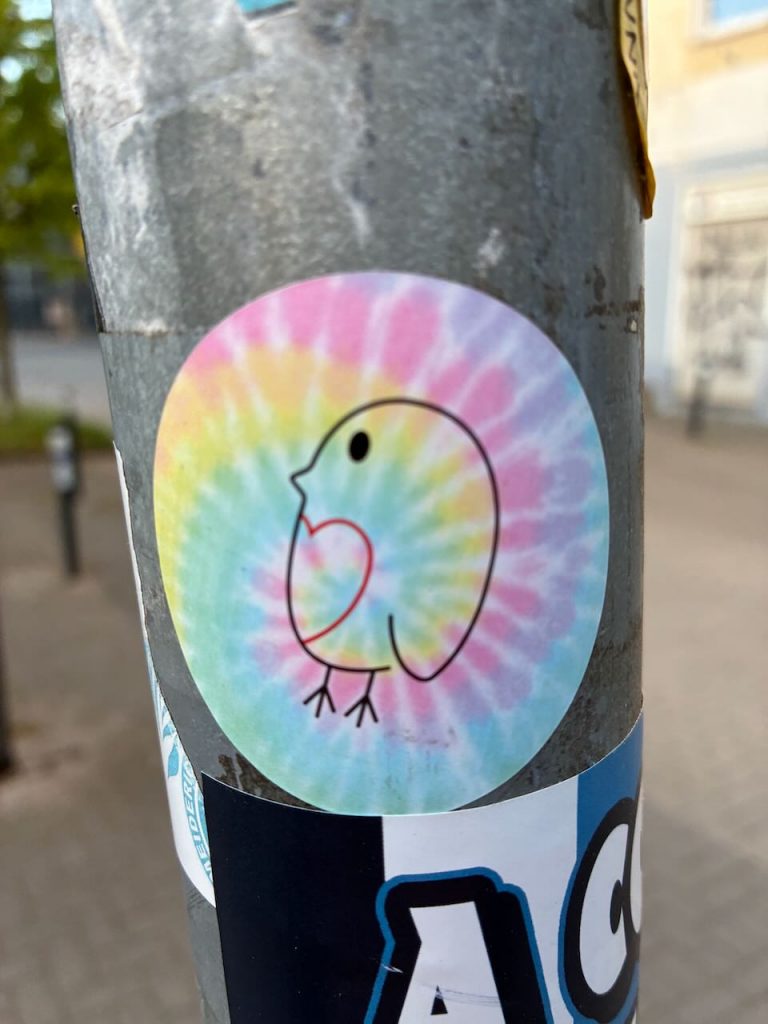 Ein kreisförmiger Aufkleber zeigt die schwarze Kontur eines kleinen Vogels vor einem pastellfarbenen Batik­hintergrund. Der Vogel ist stilisiert, hat einen runden Kopf, kurze Beine und eine herzförmige Brustlinie.