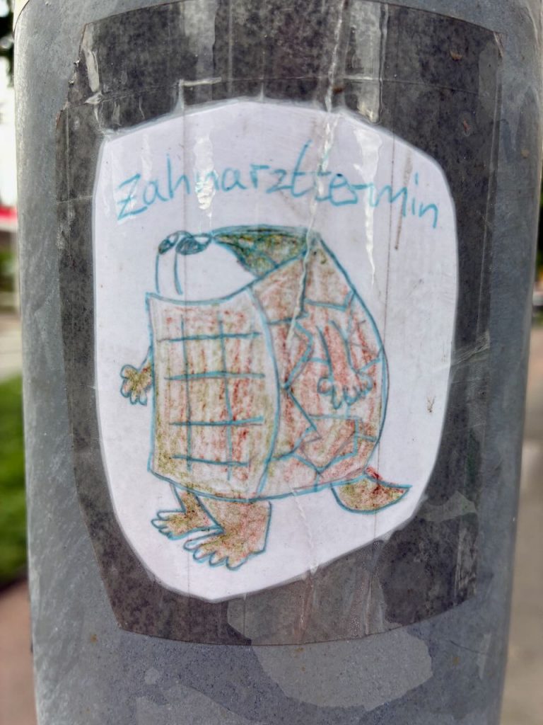 Ein handgezeichneter Schildkrötensticker auf weißem Grund. Die Schildkröte steht auf zwei Beinen, trägt ein Rechteckmuster auf dem Panzer, kleine Arme und einen länglichen Kopf. Darüber steht in türkisblauer Schrift „Zahnarzthelmin“.
