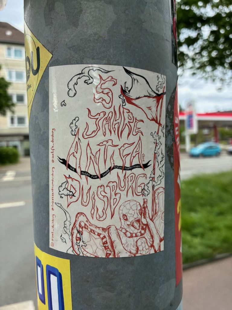 Ein rechteckiger Aufkleber mit schwarz-roter Zeichnung. Oben steht „5 Jahre Antifa Duisburg“ in flammender Schrift, unten ist eine Figur mit Maske und Tentakeln skizziert, umgeben von rauchenden Elementen.