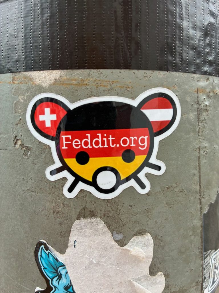 Ein konturierter Maus-Sticker mit schwarz-rotem-goldenem Deutschland-Design. Die Maus hat runde Ohren, einen runden Körper im Deutschlandfarben­muster und weiße Augenpunkte. In der Mitte steht „Feddit.org“.