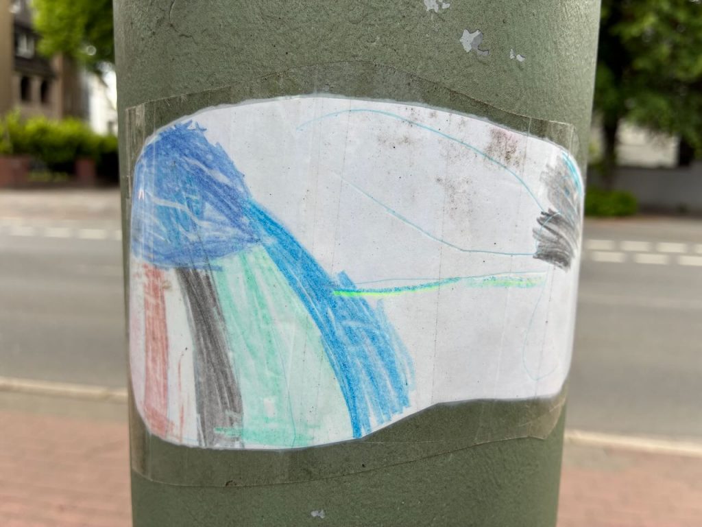 Ein ovaler Aufkleber zeigt ein Kindermotiv mit vertikalen Farbstreifen in Blau, Grün, Schwarz und Rot. Die Streifen verlaufen von unten nach oben, rechts oben ist ein grauer Bereich.