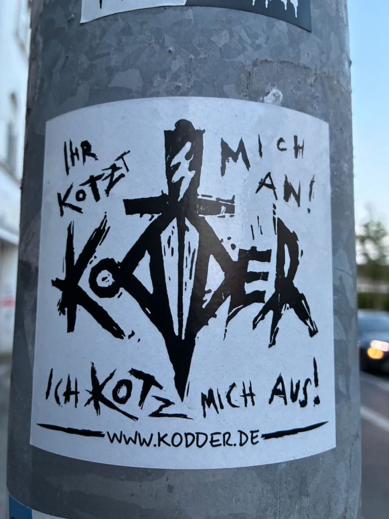 Auf einem Metallpfosten ist ein weißer rechteckiger Sticker mit schwarzer, zackiger Schrift und einem zentralen Symbol abgebildet. Oben steht „Ihr kotzt mich an!“, in der Mitte ein spitzwinkliges, geometrisches Logo und unten „Ich kotze mich aus.“ sowie darunter „www.kodder.de“.
