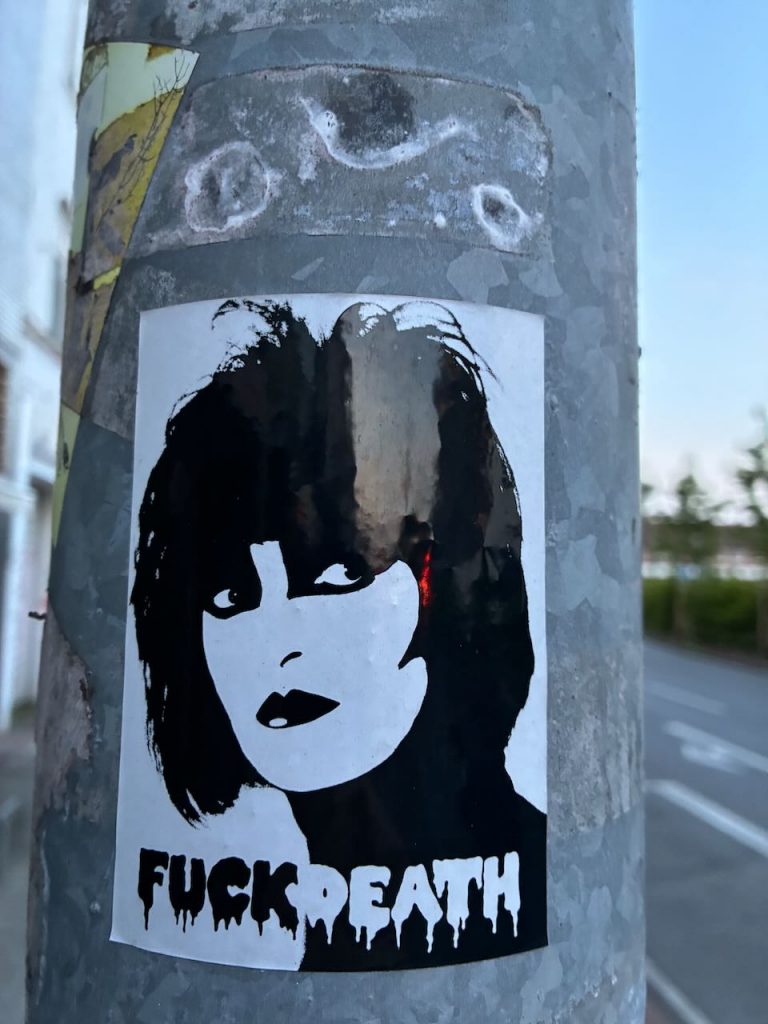Ein schwarz-weißes Sticker-Porträt zeigt eine Frau mit dunklen Haaren und Lippen, der Blick nach rechts gerichtet. Darunter steht in tropfender Schrift „Fuck Death“.
