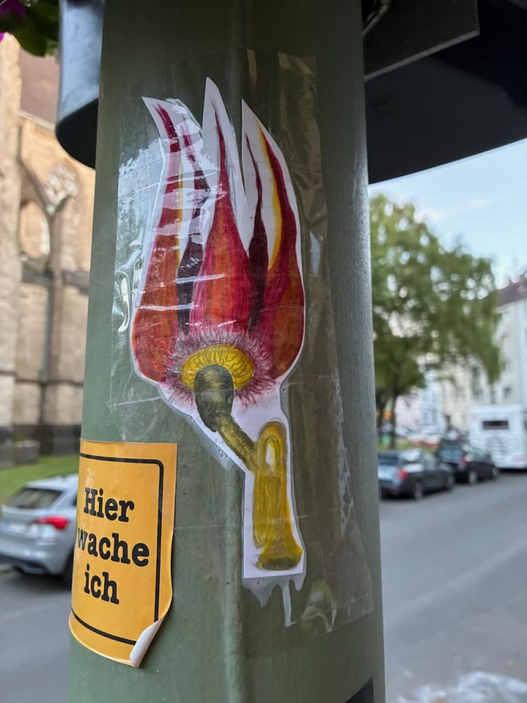 Ein bunter Sticker zeigt ein gebogenes Horn, aus dessen Trichter rote und weiße Flammen emporlodern. Der Sticker ist auf transparentem Klebeband auf einem grünen Pfosten befestigt.
