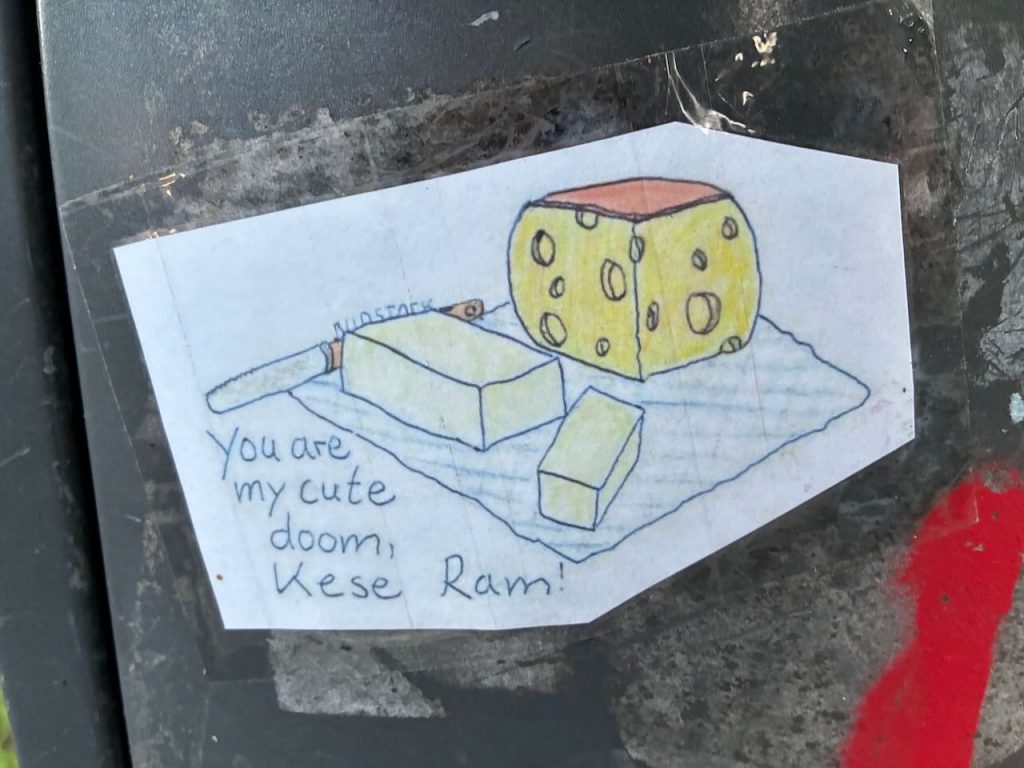 Auf weißem Hintergrund ist ein handgezeichneter gelber Würfelkäse mit Löchern zu sehen, daneben ein Butterstück und ein Messer. Darunter steht „You are my cute doom, Kese Ram!“.
