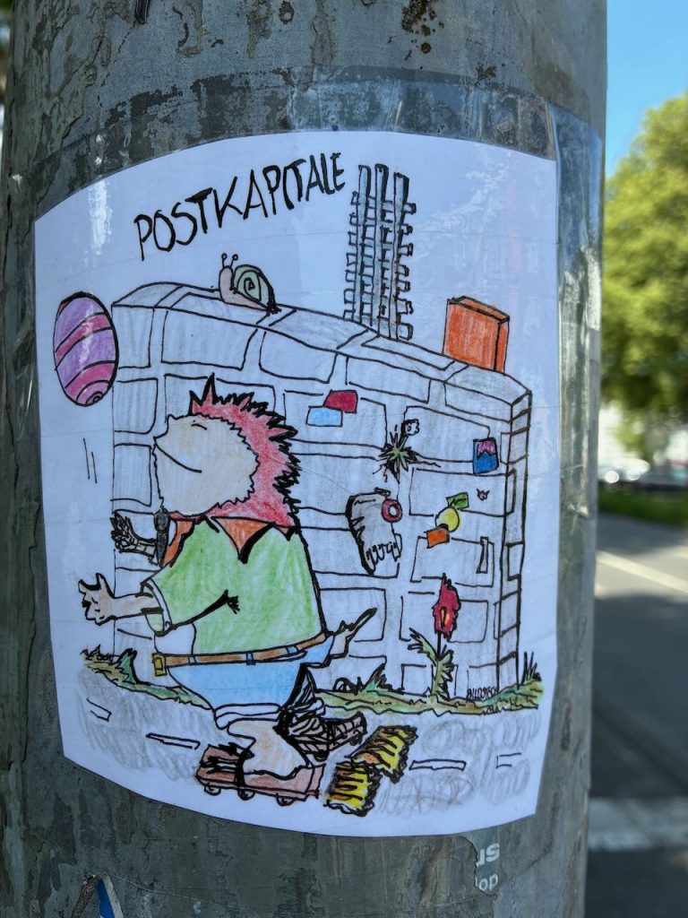 Ein gezeichneter Igel mit Skateboard läuft vor einem Postkasten entlang. Die Zeichnung ist koloriert, über dem Postkasten steht „POSTKAPSALE“.
