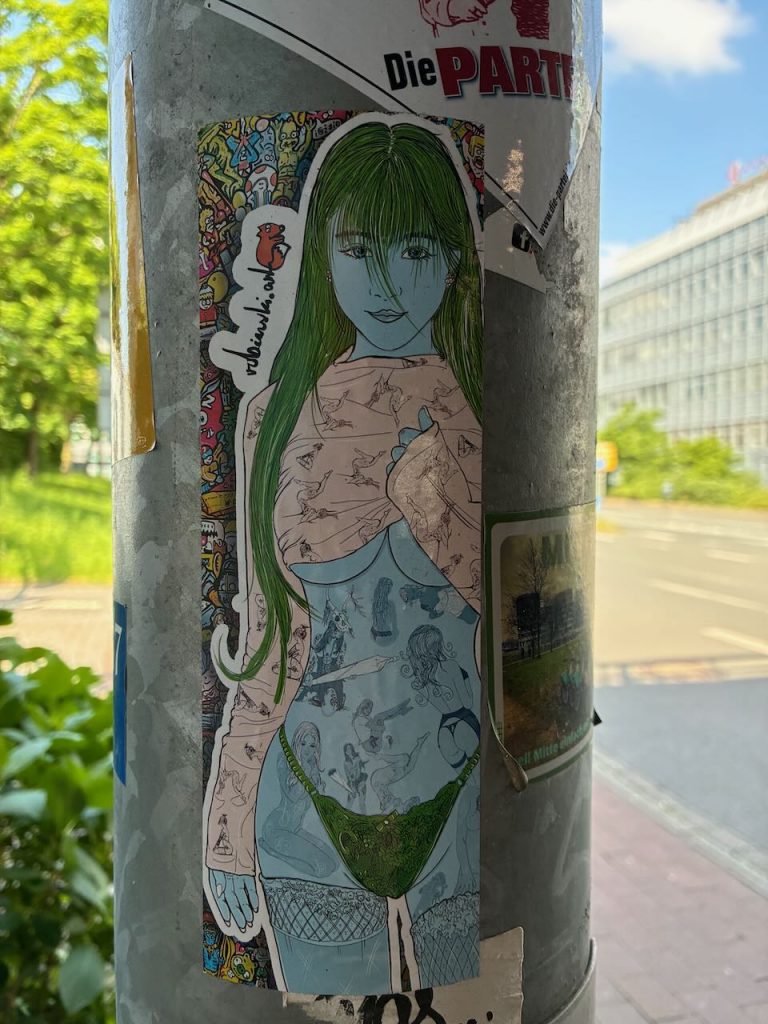 Ein hochformatiger Sticker zeigt eine blaugrüne, langhaarige Anime-Frau in grüner Unterwäsche und langärmligem Oberteil mit Mustern. Im Hintergrund sind bunte Muster erkennbar.
