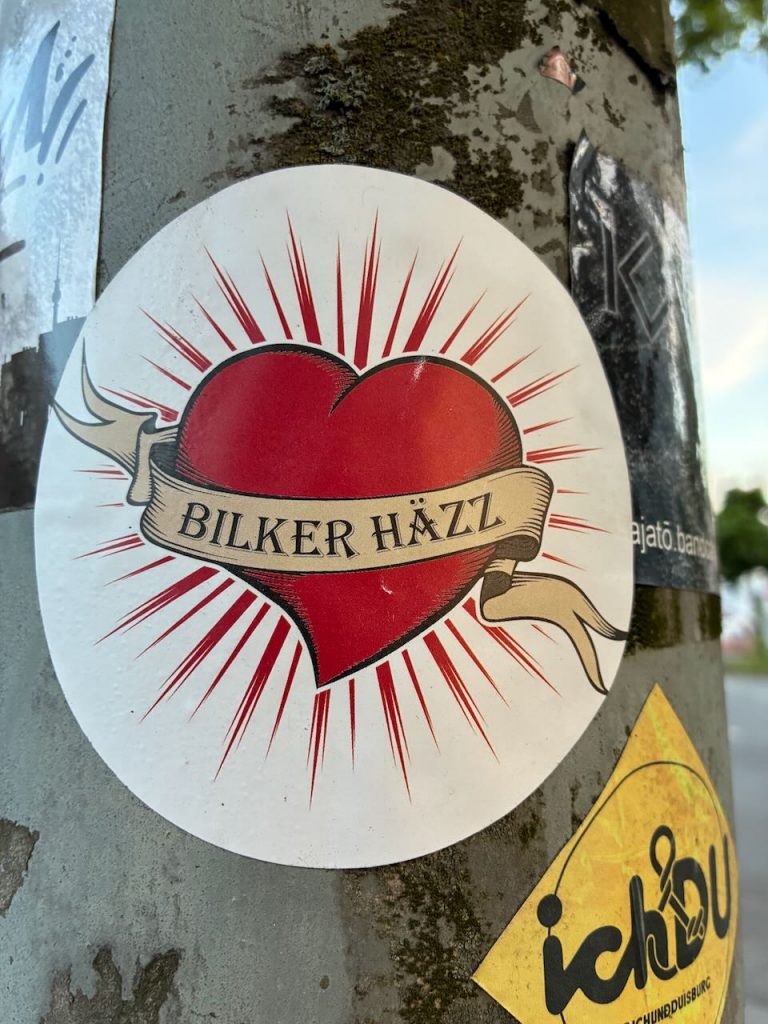 Rundes Stickerbild mit rotem Herz in der Mitte, das von goldenen Strahlen umgeben ist. Um das Herz liegt ein beige-gelbes Band mit der schwarzen Aufschrift „BILKER HÄZZ“. Das Aufkleberdesign erinnert an klassische Tattoo-Motive.
