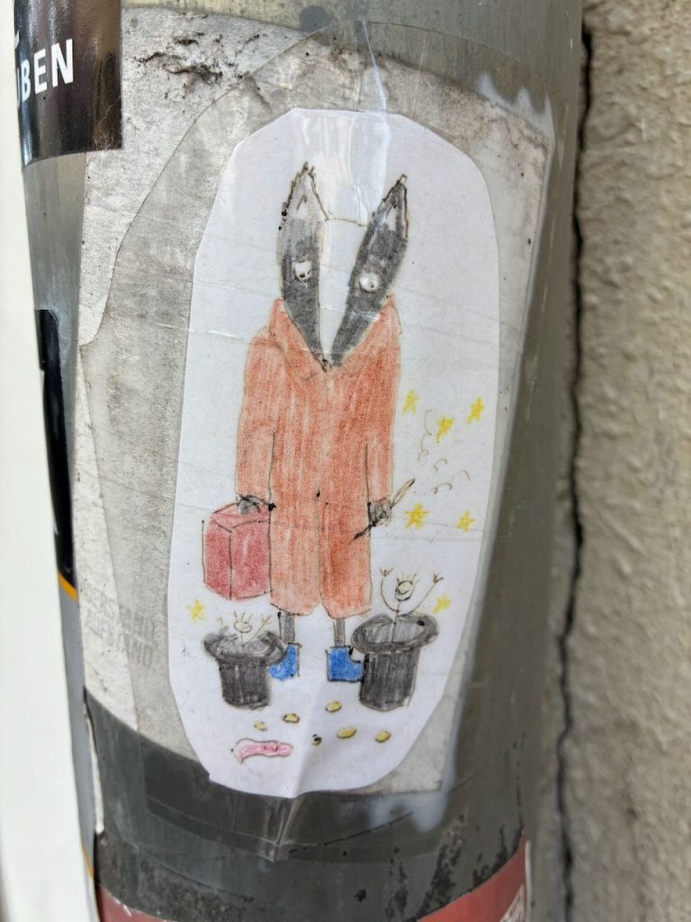 Bunt gezeichneter Aufkleber zeigt eine Figur mit grauem Dachskopf, großem roten Mantel, blauer Schuhe und Koffer in einer Hand. Zu Füßen stehen zwei lächelnde Mülleimer und einige gelbe Objekte sowie ein rosafarbenes Würmchen.
