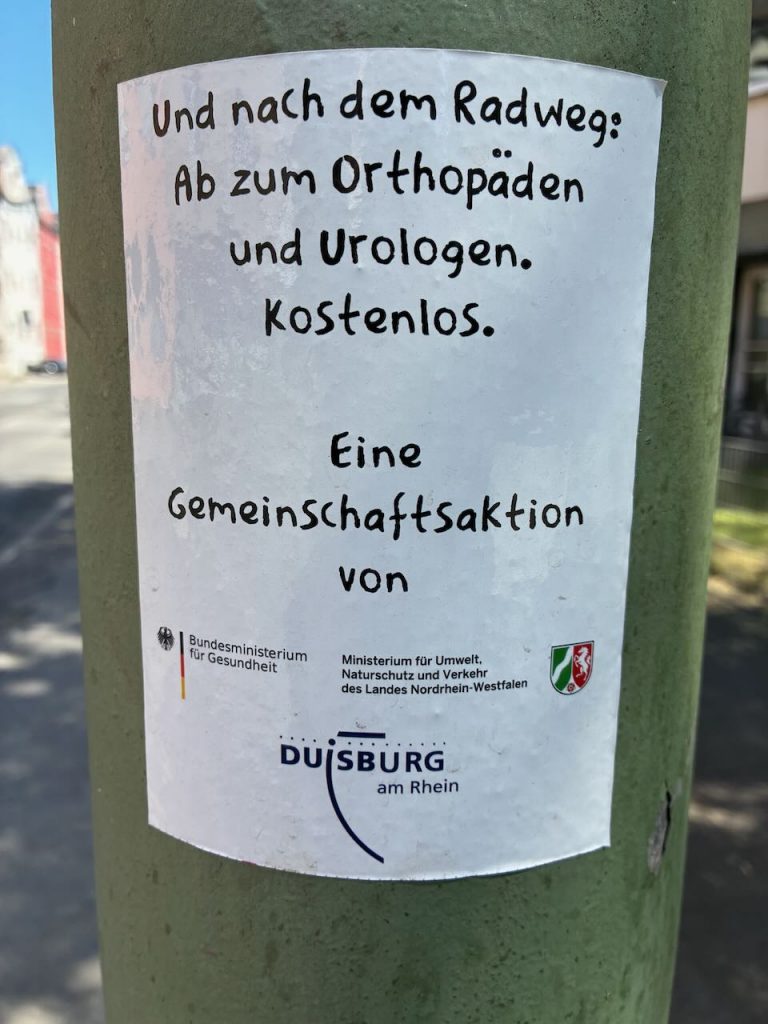 Weißer Aufkleber mit schwarzer Schrift: „Und nach dem Radweg: Ab zum Orthopäden und Urologen. Kostenlos. Eine Gemeinschaftsaktion von…“ Dazu Logos von Bundesministerien, Duisburg und NRW.
