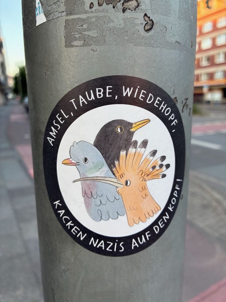 Runder Sticker mit schwarzem Rand und weißem Grund. In der Mitte gemalte Köpfe einer Amsel, Taube und eines Wiedehopfes nebeneinander. Umschrift: „AMSEL, TAUBE, WIEDEHOFP, KACKEN NAZIS AUF DEN KOPF!“
