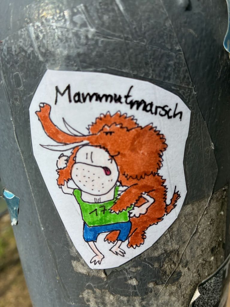 Handgezeichneter Sticker mit einer Person in grüner Startnummer, die ein kleines braunes Mammut Huckepack trägt. Darüber steht das Wort „Mammutmarsch“ handschriftlich geschrieben.
