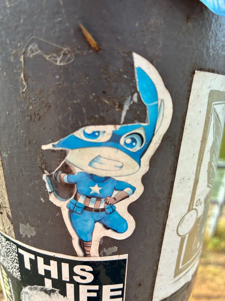 Sticker mit einer comichaften Figur von Captain America im blauen Kostüm, weißem Stern auf der Brust und großem Kopf. Die Figur hält den Schild und ist leicht schräg nach vorne dargestellt.
