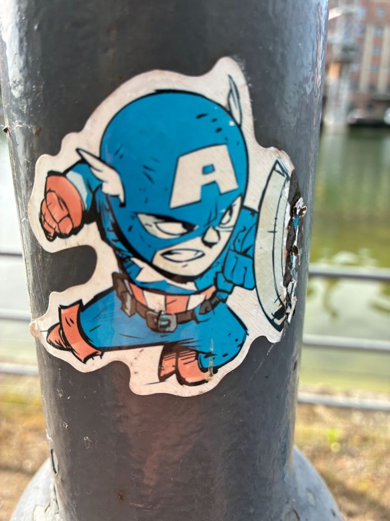 Sticker mit einer Comiczeichnung von Captain America im blauen Anzug mit großem „A“ auf der Stirn, Schild am linken Arm, der heldenhaft nach vorne springt.
