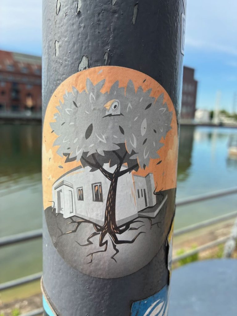Runder Sticker mit einem großen Baum, in dessen Krone ein kleiner Vogelkopf herausschaut. Unter dem Baum steht ein weißes Haus mit Fenstern und Tür, darüber ein orangefarbener Hintergrund.
