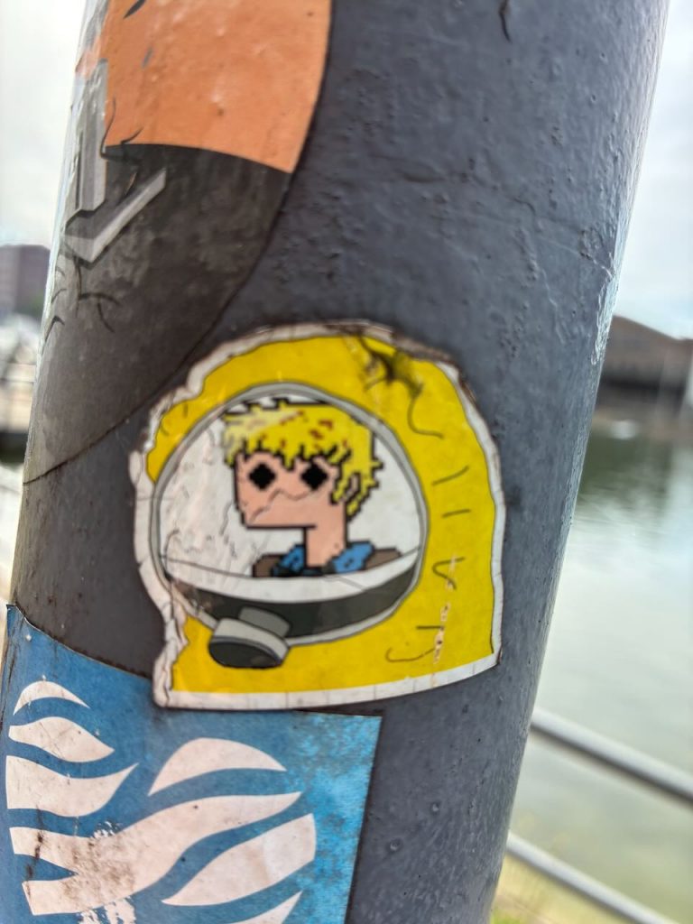 Sticker im Pixelstil, zeigt einen blonden Menschen mit Sonnenbrille, der aus einem Bullauge eines grauen Fahrzeugs oder Bootes schaut. Gelber Hintergrund.
