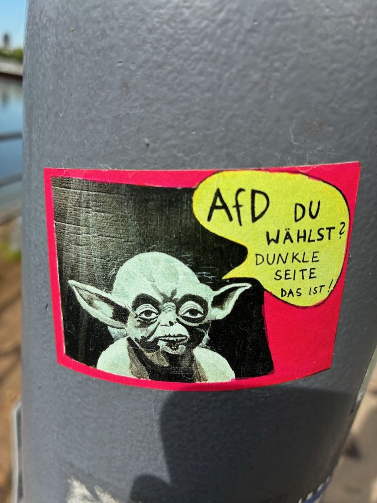 Rechteckiger Sticker mit Yoda aus Star Wars. In einer gelben Sprechblase steht: „AfD du wählst? Dunkle Seite das ist!“ Rosaroter Rahmen, schwarzer Hintergrund.

