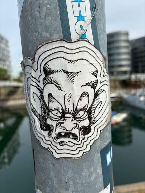 Weißer Aufkleber auf grauer Betonsäule zeigt ein gezeichnetes dämonisches Gesicht mit spitzen Ohren, langen Haaren, großen Augen, breitem Grinsen und Zähnen. Das Gesicht ist von wellenförmigen Linien umgeben.