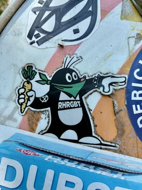 Aufgeklebter Comic-Maulwurf in Schwarz-Weiß, mit grünem Tuch über Mund und Nase. Linke Hand hält eine Karotte, rechte zeigt nach rechts. Auf dem Bauch steht „RHRGBT“. Der Sticker sitzt auf abgenutzter, teils rostfarbener Oberfläche mit weiteren Aufklebern.