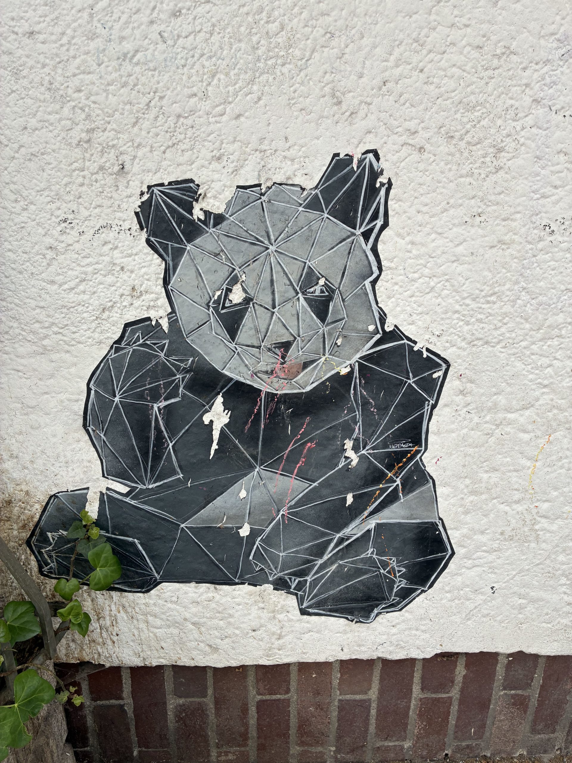 Großer Sticker an einer hellen Hauswand über einem Ziegelsockel. Dargestellt ist ein sitzender Panda in dunklen Grau- und Schwarztönen. Sein Körper besteht aus vielen polygonalen Flächen mit feinen Linien, die an ein 3D-Drahtgitter erinnern. Teile des Stickers sind leicht abgerissen.