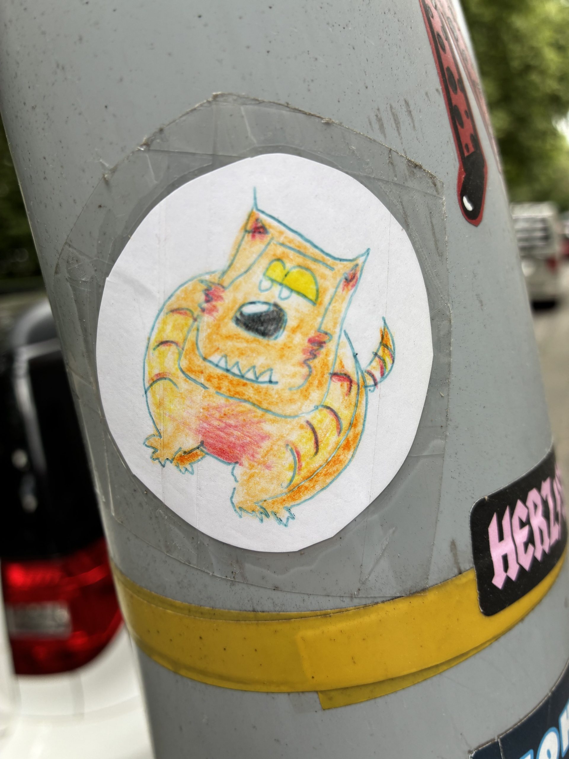 Runder Sticker auf einem grauen Mast, mit durchsichtigem Klebeband befestigt. Darauf ein handgezeichnete, orange-gelber Tiger mit gelben Augen, schwarzer Nase und spitzen Zähnen. Das Tier hat kurze Beine, kleine Ohren und einen gestreiften Schwanz. Die Zeichnung wirkt kindlich und mit Buntstiften koloriert.